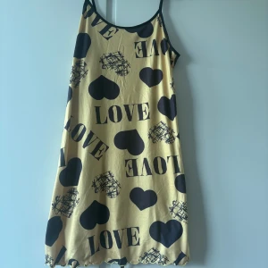 Gul kortklänning med hjärtan och text - Snygg gul kortklänning med svarta hjärtan och texten LOVE tryckt över hela tyget. Klänningen har tunna axelband och en avslappnad passform, perfekt för varma dagar. Materialet känns mjukt och lätt mot huden.