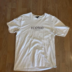 Vit t-shirt från Icono Couture S - Vit t-shirt från Icono Couture med tryck framtill och en stor grafisk design på ryggen med statyhuvuden och dödskalle i färg. Modellen är relaxed fit och har klassisk rund hals och korta ärmar. 