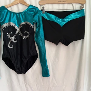 Grön och svart gymnastikdräkt + kort byxa - Säljer ett set med en glansig gymnastikdräkt i grön och svart med långa ärmar, samt ett par korta matchande byxor. Tröjan har silvriga detaljer och paljetter framtill som ger en snygg look. Materialet är stretchigt och passar perfekt för träning eller dans. Storlek saknas på plaggen, men jag chansar på att de passar s/m