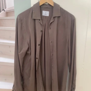 Brun långärmad skjorta från Bläck - Stilren brun skjorta från Bläck med klassisk krage och knäppning framtill. Skjortan är långärmad och har en avslappnad passform, perfekt att bära öppen eller stängd. Tillverkad i mjukt material som känns skönt mot huden.