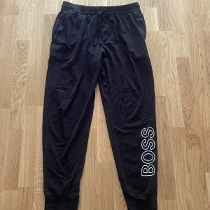 Svarta mjukisbyxor från Hugo Boss - Svarta mjukisbyxor från Hugo Boss med stor vit BOSS-logga längs ena benet. Byxorna har resår och snörning i midjan samt muddar vid bensluten. Perfekta för chill eller träning, tillverkade i mjukt material för maximal komfort.