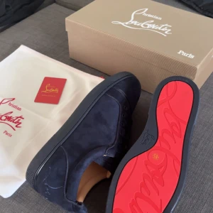 Mörkblå sneakers från Christian Louboutin - Exklusiva mörkblå sneakers från Christian Louboutin i mocka med den ikoniska röda sulan. Skorna har rund tå, snörning och en stilren design som sticker ut. Perfekta för dig som vill ha något unikt och lyxigt i garderoben.
