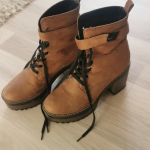 Bruna mockaboots med klack och snörning - Snygga bruna boots i mocka med kraftig klack och plattform. Skorna har svarta snören, grov sula och dekorativ rem med spänne upptill. Perfekta för dig som vill ha en cool och trendig look med extra höjd. Storlek 40 normal i storleken. 