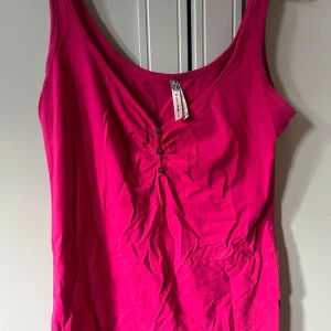 Rosa linnetopp från Bershka - Snygg rosa linnetopp från Bershka med v-ringning och små knappar framtill. Toppen har en lätt rynkad detalj vid bysten och är ärmlös, perfekt för en trendig och färgstark look. Mjuk och skön i bomullsmaterial