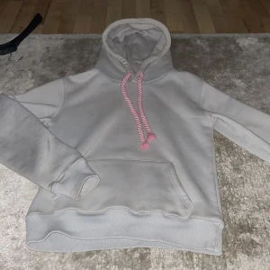 Ljusgrå t synaworldtracksuit - Ljusgrå hoodie från Synaworld med fet rosa graffiti-tryck på huvan och matchande rosa snören. Klassisk känguruficka framtill och mjuk bomullskänsla. Perfekt för dig som gillar streetstyle och vill sticka ut med coola detaljer. storlek S 