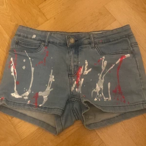 Pepperts jeansshorts med färgstänk - Snygga ljusblå jeansshorts från Pepperts i super skinny fit. Shortsen har coola röda och vita färgstänk framtill och baktill, klassiska fickor och bälteshällor. Perfekta för dig som vill sticka ut med en kreativ och trendig look i sommar.
