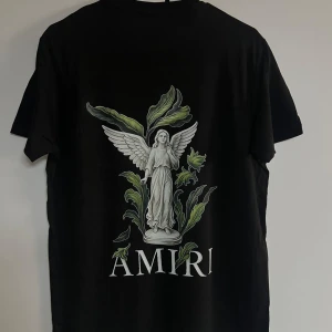 Svart Amiri t-shirt med ängeltryck - Svart t-shirt från Amiri med stort tryck av en vit ängel omgiven av gröna blad på ryggen och mindre tryck på bröstet. Klassisk rund hals och korta ärmar. Snygg och stilren design med exklusiv känsla.