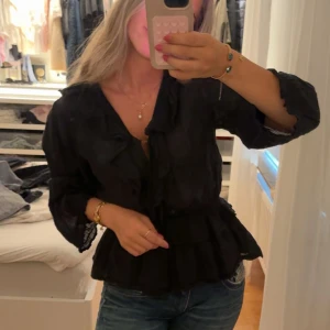 Vardags blus från neo noir - Elegant svart blus i lätt, transparent tyg med subtilt mönster. Blusen har volangdetaljer längs knäppningen och vid ärmslut samt en markerad midja.