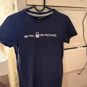 Mörkblå Sail Racing t-shirt XS - Snygg mörkblå t-shirt från Sail Racing i 100% bomull. Klassisk passform med korta ärmar och rund hals. Framsidan har vit Sail Racing-logga och en liten logga finns även på ryggen. Perfekt för dig som gillar stilren och sportig look.