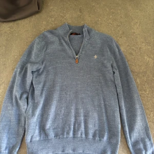 Blå stickad half zip-tröja Morris - Säljer en blå stickad tröja från Morris med half zip och ribbade muddar. Tröjan har en liten broderad logga på bröstet och hög krage. Perfekt för dig som gillar stilren och klassisk look. Passar till jeans eller chinos.