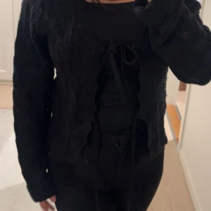 Svart stickad kofta med snörning - Säljer en svart stickad kofta med snyggt hålmönster och knytning framtill. Koftan har långärmade ärmar och en lite croppad passform. Perfekt att slänga över en topp för en chill men ändå stilren look.