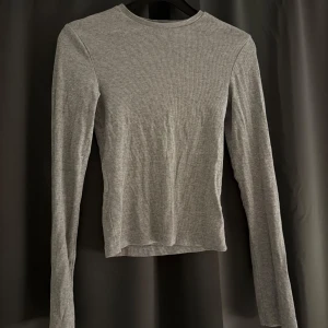 Grå ribbad långärmad tröja - En enkel och stilren grå långärmad tröja med ribbad struktur. Tröjan har en rund halsringning och är figurnära i modellen, perfekt att matcha med jeans eller kjol. Mjuk och skön, passar året runt.
