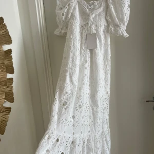 Vit långklänning med broderade detaljer - Superfin vit långklänning med broderade hålmönster och puffiga korta ärmar. Klänningen har smock upptill, knytning vid bysten och volang nertill. Perfekt för sommarens alla tillfällen! Nypris 2199