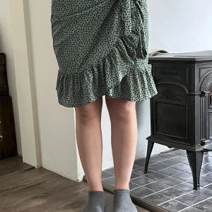 Grön blommig omlottkjol med volang - Supersöt grön kortkjol med svarta blommor och omlottmodell. Kjolen har en volangkant nertill och knyts i sidan med en rosett. Perfekt för dig som gillar en lekfull och trendig stil. Lätt och skönt material som passar till många olika outfits.