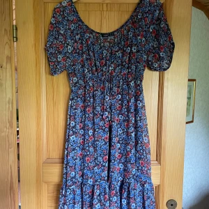Blommig långklänning  - Supersöt långklänning från Lindex med blå, röda och beige blommor på mörk botten. Klänningen har korta puffärmar, resår i midjan och volang nertill. Perfekt för sommarens alla tillfällen och ger en romantisk vibe.