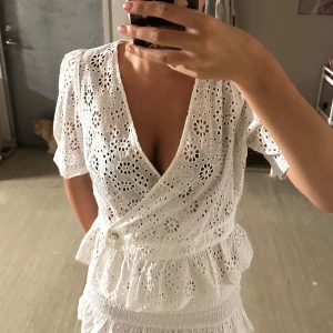 Vit broderad omlottblus med pärlknapp - Superfin vit omlottblus i bomull med broderade blommor och hålmönster. Blusen har korta ärmar, volang nertill och en snygg pärlknapp i midjan. Perfekt för sommarens alla tillfällen och ger en romantisk vibe.