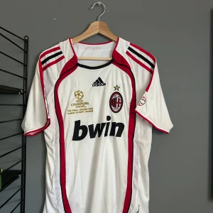 AC Milan Kaka' 22 Champions League tröja - Säljer en ikonisk AC Milan bortatröja från Champions League-finalen 2007 med Kaka' och nummer 22 på ryggen. Tröjan är vit med röda och svarta detaljer, Adidas-logga, klubbmärke och Bwin som sponsor. Klassisk passform i lätt och ventilerande material. Helt ny med tagg en replika