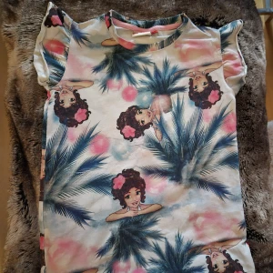 Färgglad t-shirt från LEGO Wear - Superfin t-shirt från LEGO Wear med färgglatt tryck av tecknade tjejer, palmer och rosa moln. T-shirten har korta ärmar med lätt volang och rund halsringning. Mjukt material som känns skönt mot huden. Perfekt för dig som gillar unika prints och lekfull stil.