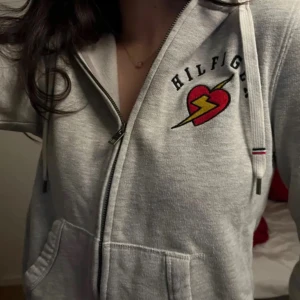 Grå hoodiezip Tommy Hilfiger - Grå hoodiejacka från Tommy Hilfiger med dragkedja, huva och snörning. På bröstet finns ett broderat rött hjärta med blixt och svart text. 