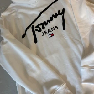Vit hoodie från Tommy Jeans - Säljer en snygg vit hoodie från Tommy Jeans med svart logga och klassisk flagga på ryggen. Helt ny med prislapp kvar