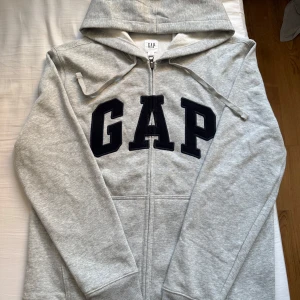 Grå GAP hoodie med dragkedja - Snygg ljusgrå hoodie från GAP med stor svart logga framtill. Tröjan har huva med snörning, dragkedja hela vägen och långa ärmar. Helt oanvänd 
