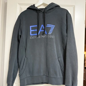 Svart EA7 Emporio Armani hoodie - Snygg svart hoodie från EA7 Emporio Armani med blå logga framtill och tryck längs ryggen. Tröjan har huva med snörning, känguruficka och långa ärmar. Perfekt för dig som gillar streetwear och vill sticka ut med ett exklusivt märke.