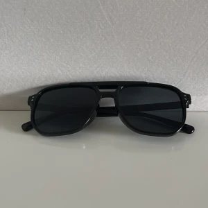 Svarta grisch solglasögon - Snygga svarta solglasögon med klassisk pilotform och mörka glas. Ramen är i blank svart plast med dubbla nitar vid bågens framkant. Perfekt accessoar för en cool look i solen.