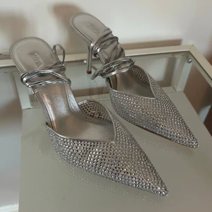 Silvriga pumps med strass från Zara - Glittriga pumps från Zara i metallic silver med spetsig tå och smal klack. Skorna har snygga remmar som snöras runt vristen och är täckta av små strass-stenar för extra bling.