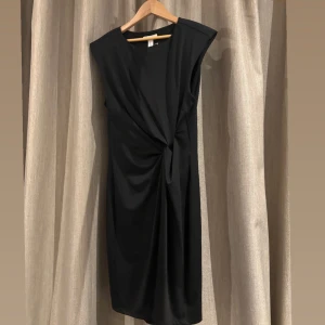 Svart klänning med knytdetalj - Elegant svart klänning med en unik knytdetalj framtill. Den är ärmlös och har en stilren design som passar perfekt för olika tillfällen. Klänningen har en smickrande passform och är tillverkad i ett mjukt material.