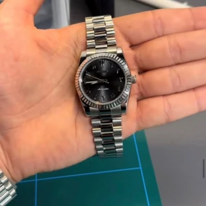 Silverfärgad klocka med svart urtavla - Elegant klocka med en silverfärgad länkarmband och en stilren svart urtavla. Klockan har en klassisk design med tydliga markeringar och en räfflad bezel. Perfekt för den som söker en tidlös accessoar.