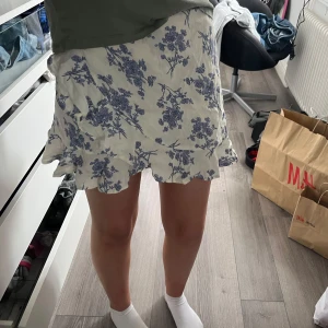 Blommig kjol med volang - Säljer en söt kortkjol med blått blommönster på vit bakgrund. Kjolen har volangdetaljer som ger en lekfull touch. Perfekt för varma dagar!