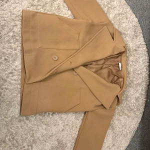 Beige kavaj från Design by Si - Snygg beige kavaj från Design by Si med klassisk krage och knappar framtill. Perfekt för en stilren look. Knappt använd 🥰