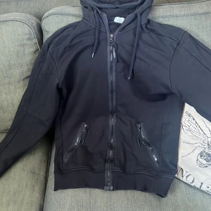 Svart hoodie från C.P. Company - Säljer en svart hoodie från C.P. Company med dragkedja och huva. Den har unika glasögon integrerade i huvan och snörning för justering. Perfekt för en cool och avslappnad stil.