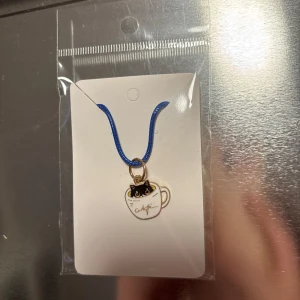 Halsband med katt i kopp - Charmigt halsband med en söt berlock föreställande en svart katt i en vit kopp. Berlocken hänger på ett blått band och ger en lekfull touch till din stil.