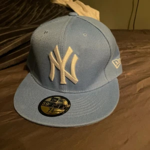 Ljusblå keps från New Era - Snygg ljusblå keps från New Era med vit broderad logga framtill. Kepsen har en klassisk 59FIFTY design med en grå undersida på skärmen. Perfekt för att ge din outfit en sportig touch.