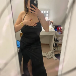 Svart långklänning med slits - Elegant svart långklänning med smala axelband och en stilren slits framtill. Klänningen har en åtsittande passform och en öppen rygg som ger en sofistikerad look. Perfekt för festliga tillfällen. Jag har knutit bandet då den gick sönder men de ser ingen fara ut 