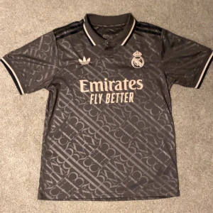 Grå fotbollströja från Adidas - Säljer en grå fotbollströja från Adidas med Real Madrid-logga och 'Emirates Fly Better' tryck. Tröjan har ett subtilt mönster och korta ärmar med svarta och vita detaljer på krage och ärmslut. Perfekt för fotbollsfans! ⚽