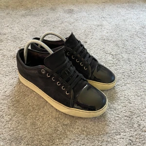 Svarta sneakers från Lanvin - Snygga svarta sneakers från Lanvin med en kombination av mocka och glansigt material på tån. Skorna har en vit sula och klassisk snörning. Perfekta för en stilren look.