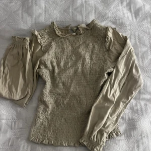 Beige blus från Zara - Säljer en söt beige blus från Zara med smockdetaljer och volanger. Blusen har långa ärmar och en fin spetskant vid halsen. Perfekt för en stilren look! 