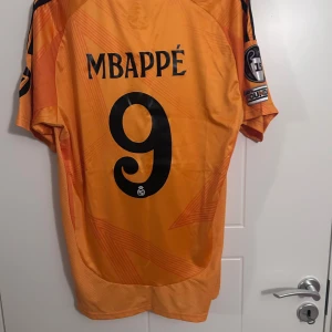 24-25 Relmadrid fotbollströja (Mbappé) - Säljer en orange fotbollströja från Adidas med svart text och detaljer. Tröjan har korta ärmar och ett tryck med 'Mbappé' och nummer 9 på baksidan. Perfekt för fotbollsfans!