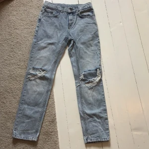 Ljusa jeans med slitningar - Säljer ett par ljusa jeans med coola slitningar på knäna. De har en klassisk femficksdesign och knappgylf. Perfekta för en avslappnad stil.