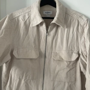 Beigevit overshirt/jacka från Strömberg - Snygg beige manchesterjacka från Strömberg i finaste bomull. Jackan har dragkedja framtill och två bröstfickor med knapplås. Perfekt för en stilren look inför utgång/finare event eller som en vardaglig elevated look. Nypris: 899kr