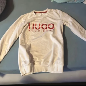 Vit tröja från Hugo Boss  - Säljer en stilren vit tröja från Hugo Boss med röd logotyp på bröstet och ärmen. Tröjan har långa ärmar och rund halsringning. Perfekt för en avslappnad och trendig look.