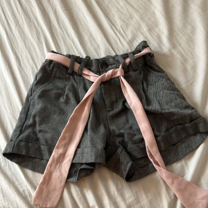 Grå shorts med rosa bälte - Snygga grå shorts med subtilt mönster och ett rosa knytbälte. Perfekta för en stilren look. De har en normal passform och är tillverkade i ett bekvämt material⭐️