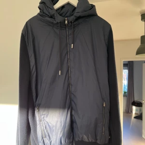 Svart jacka från Hugo Boss - Säljer en stilren svart jacka från Hugo boss  i storlek XL. Jackan har en kombination av syntetmaterial och stickade detaljer, med dragkedja och huva. Perfekt för höst och vår. Nypris 5000kr. Hål i fickan inne i jackan. Passar L också