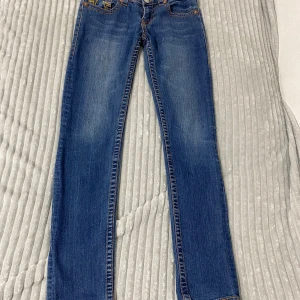Blå jeans från True Religion -  Snygga blå jeans från True Religion med ikonisk gul brodering på bakfickorna. De har en klassisk femficksdesign och en knappgylf. Perfekta för en avslappnad stil. Står ej för frakt. 