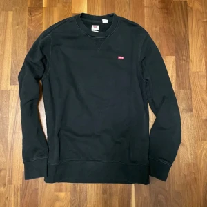 Svart tröja från Levi's - Säljer en stilren svart tröja från Levi's med rund halsringning och liten röd logga på bröstet. Perfekt för en avslappnad look. Tröjan har långa ärmar och ribbade muddar vid ärmslut och nederkant.