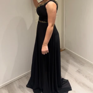 Svart balklänning med spetsdetaljer - Elegant svart balklänning med vackra spetsdetaljer vid halsringning och midja. Klänningen har en ärmlös design och en flytande kjol som ger en sofistikerad look.