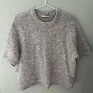 Grå stickad tröja från H&M - Säljer en mysig grå stickad tröja från H&M. Tröjan har en rund halsringning och korta ärmar, perfekt för lager på lager-stil. Den är enkel och stilren, vilket gör den lätt att matcha med olika outfits.