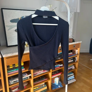 Svart ribbad omlottopp från Anna Field - Snygg svart ribbad omlottopp från Anna Field i återvunnen bomull. Toppen har en v-ringad design och långa ärmar, perfekt för en stilren look. Den omlottade stilen ger en modern touch. Passar bra till både jeans och kjol.